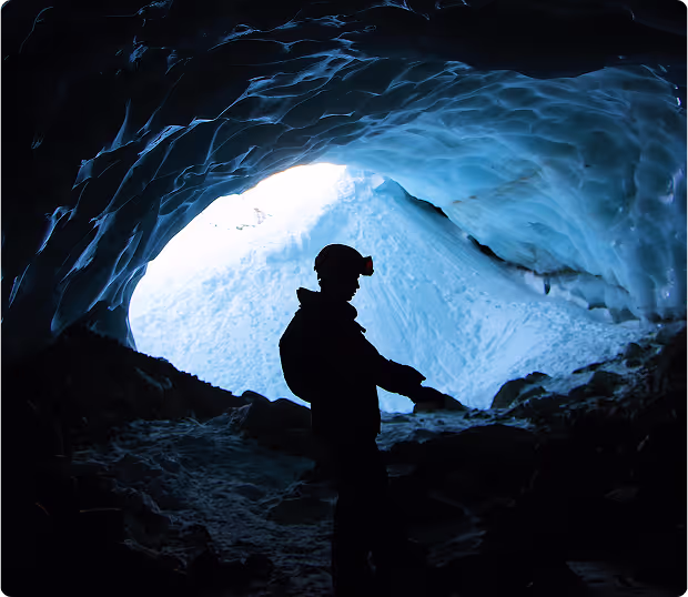 A silhouetted figure wearing a helmet with a light stands at the bright entrance of a dark mine or ice cave.